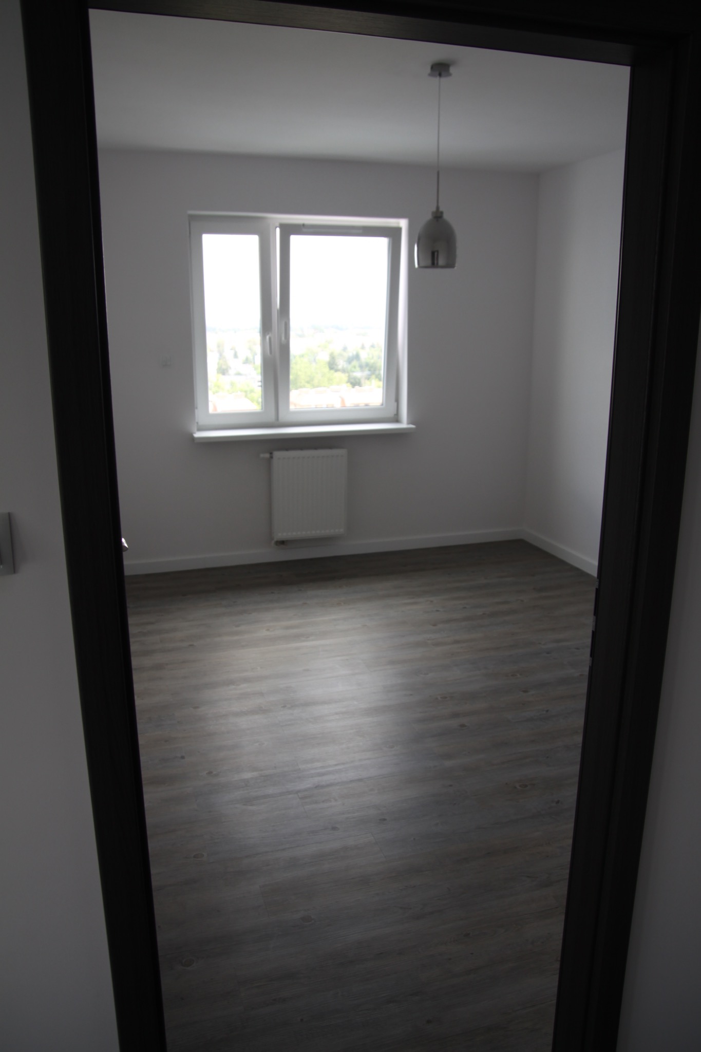 Pusty, nowocześnie wykończony pokój z panelami podłogowymi w odcieniach szarości, widok przez ciemną futrynę drzwiową, z oknem i wiszącą lampą w tle.