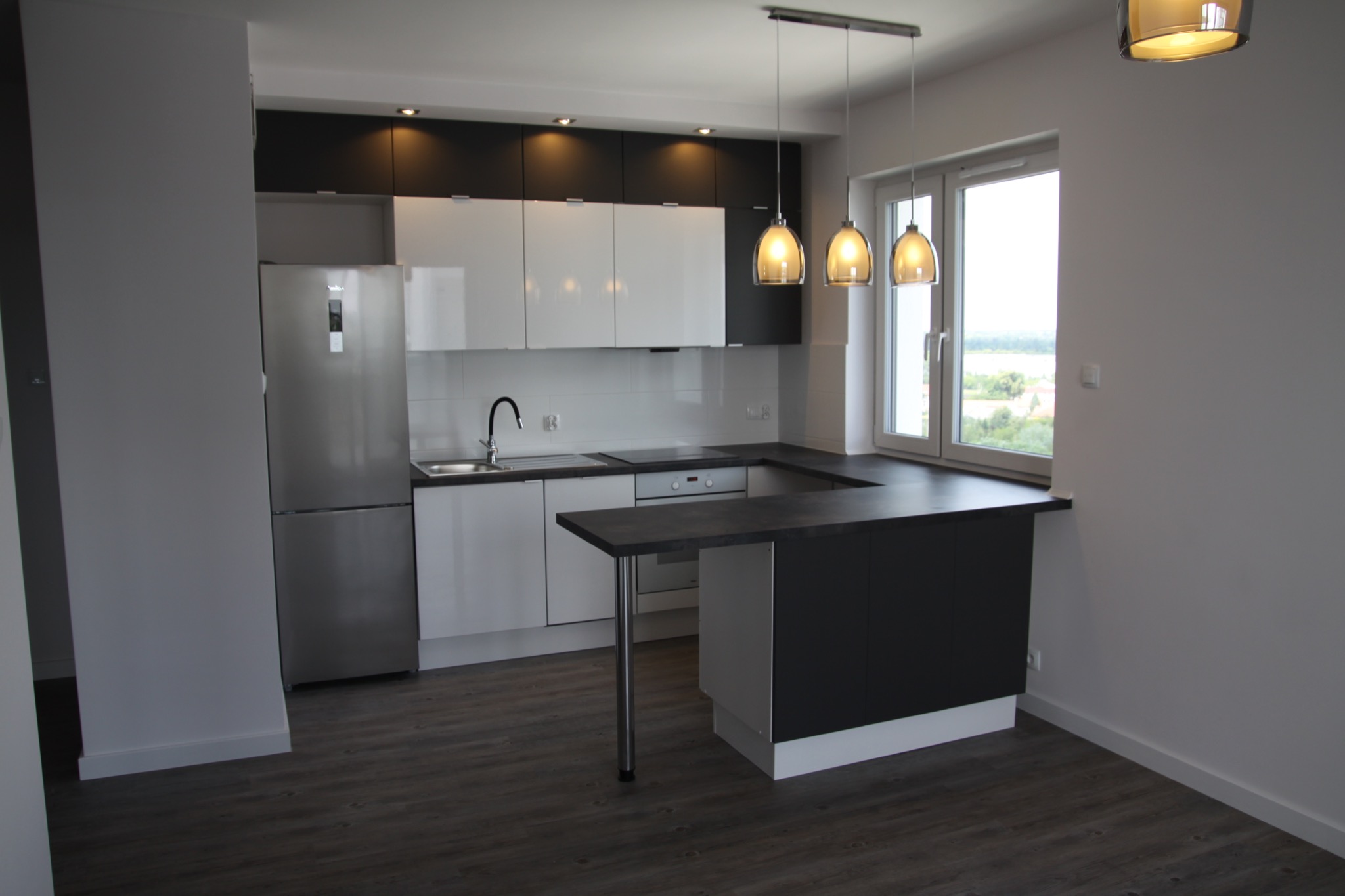 Nowoczesna kuchnia z białymi i czarnymi szafkami, stalową lodówką, barkiem z pojedynczą nogą i trzema wiszącymi lampami z bursztynowego szkła, widok na zewnątrz przez okno.