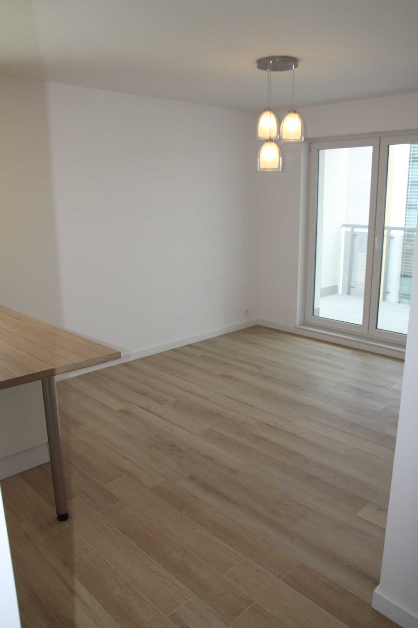 Jasne panele podłogowe imitujące drewno w pustym, nowym pomieszczeniu z białymi ścianami i potrójną lampą wiszącą, widok na balkon przez przeszklone drzwi.