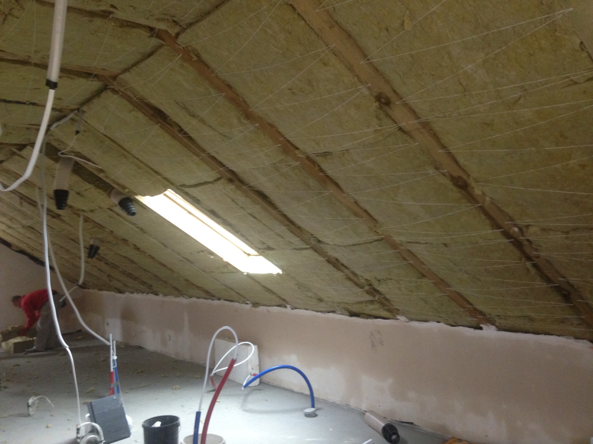 Montaż izolacji z wełny mineralnej na poddaszu, widoczne drewniane krokwie i siatka podtrzymująca, w tle pracownik w czerwonej koszulce umieszcza materiał izolacyjny.