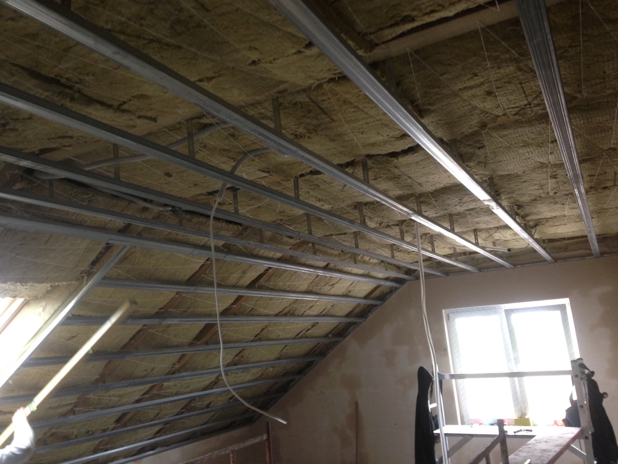 Poddasze w trakcie ocieplania wełną mineralną, widoczne metalowe profile konstrukcyjne pod płytę karton-gips, okno dachowe i fragment ściany szczytowej.