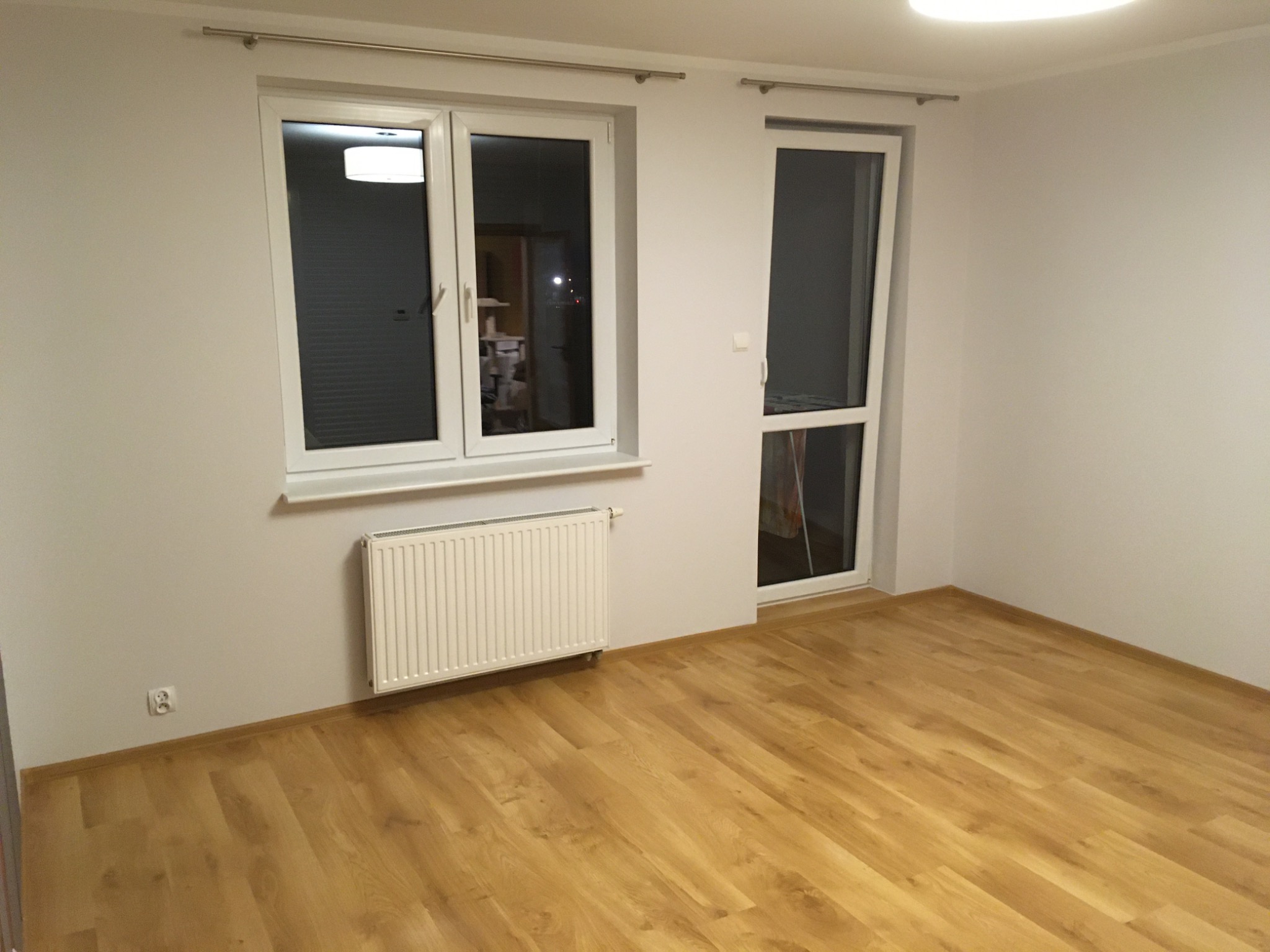 Pusty pokój z panelami podłogowymi w odcieniach ciepłego drewna, białymi ścianami, grzejnikiem pod oknem i drzwiami balkonowymi.