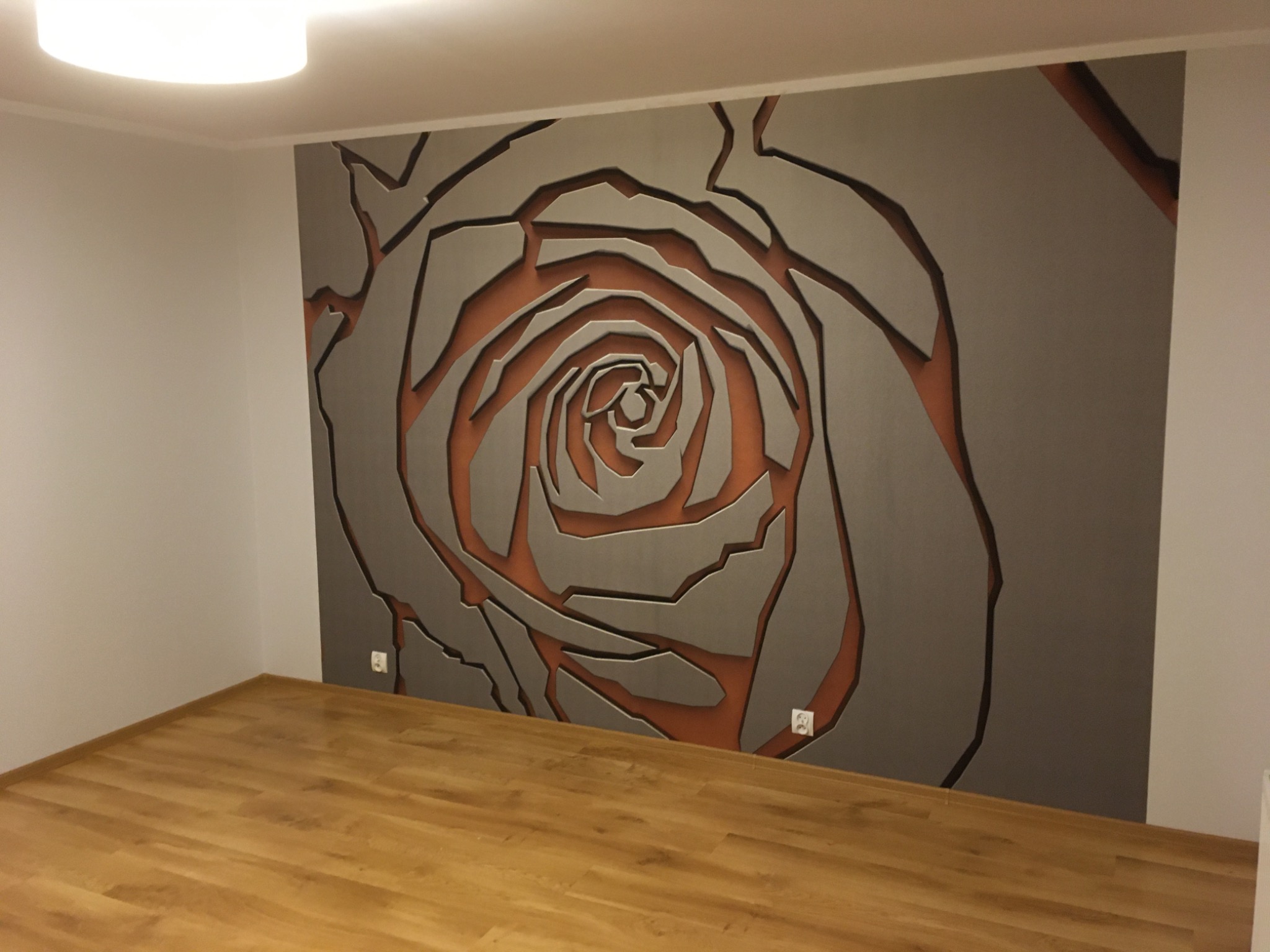 Ściana z dekoracyjną, trójwymiarową aplikacją w kształcie róży, szaro-pomarańczowa kolorystyka, drewniana podłoga, minimalistyczne wnętrze.