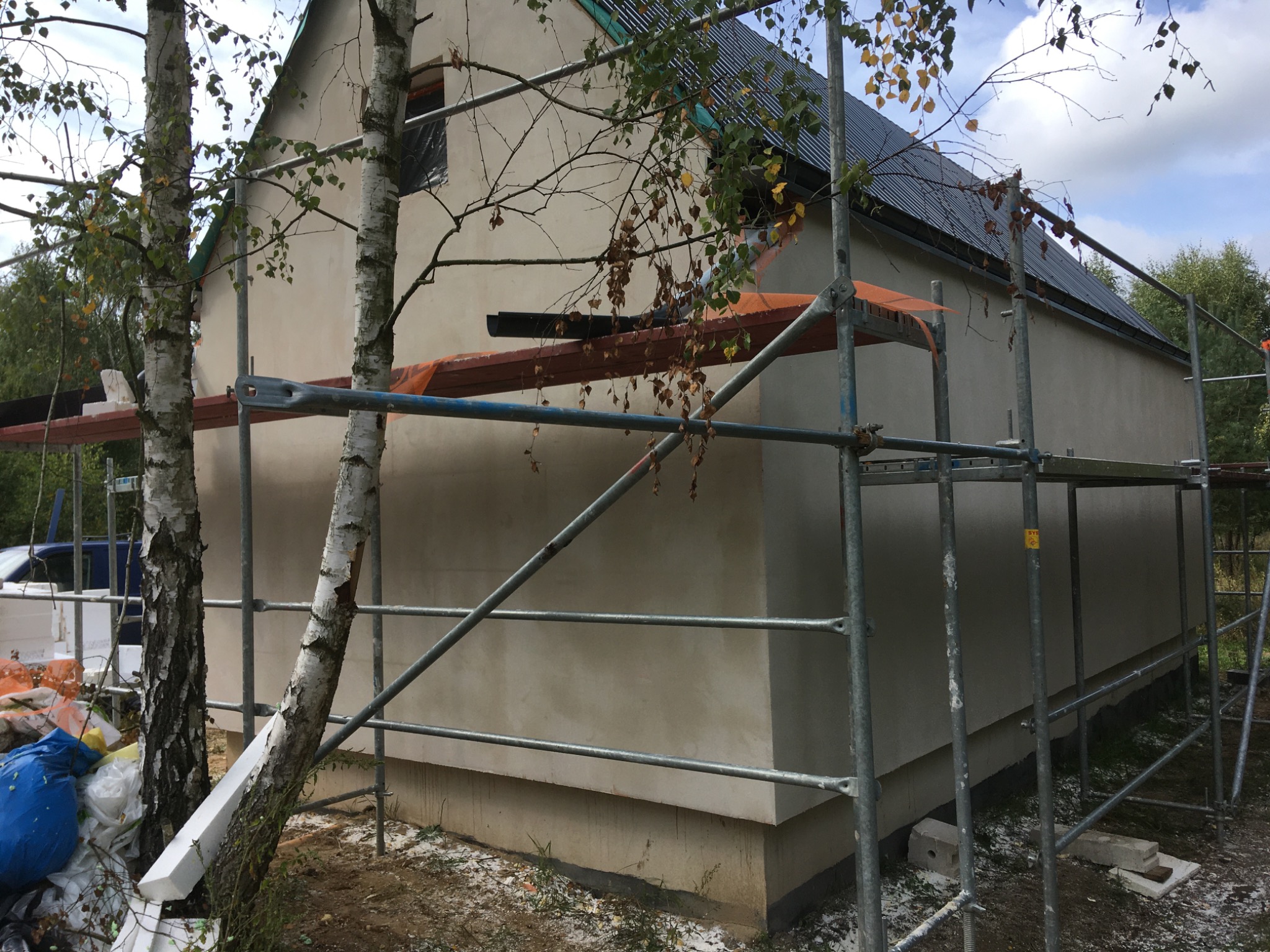 Budynek jednorodzinny w trakcie prac wykończeniowych elewacji, widoczne rusztowania, świeżo nałożony tynk, fragment dachu z blachy oraz brzozy na pierwszym planie.