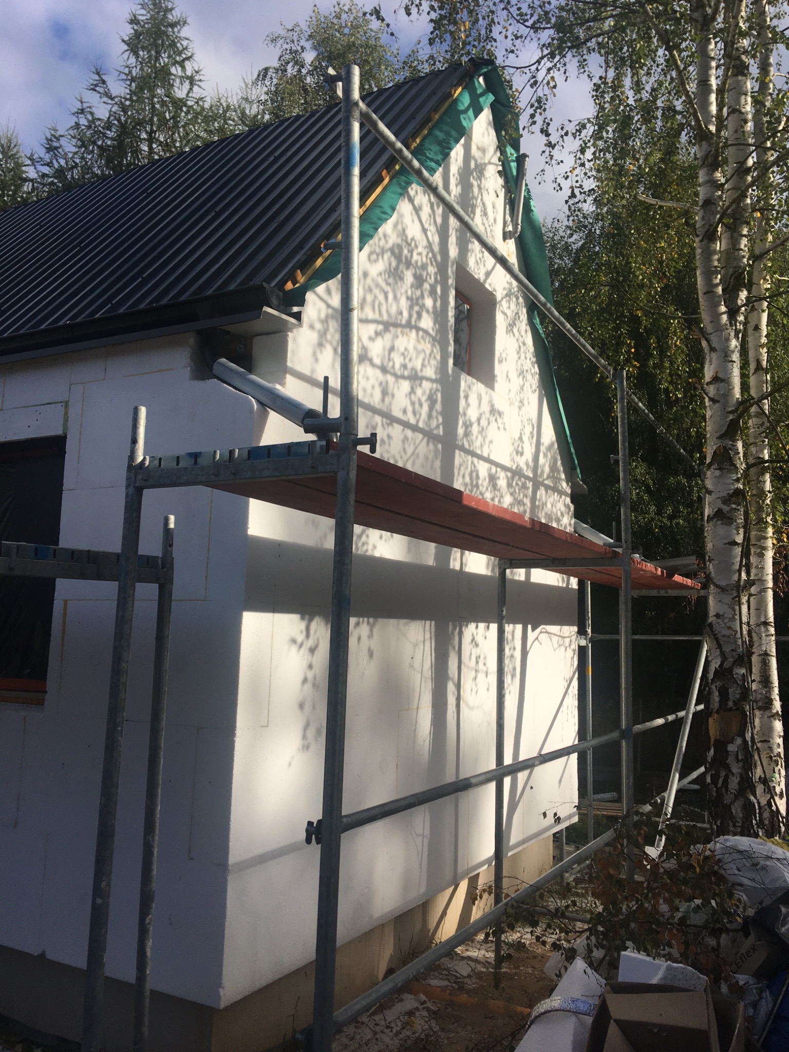 Budynek w trakcie ocieplania styropianem, widoczne rusztowanie, elementy ocieplenia i drzewa w tle.