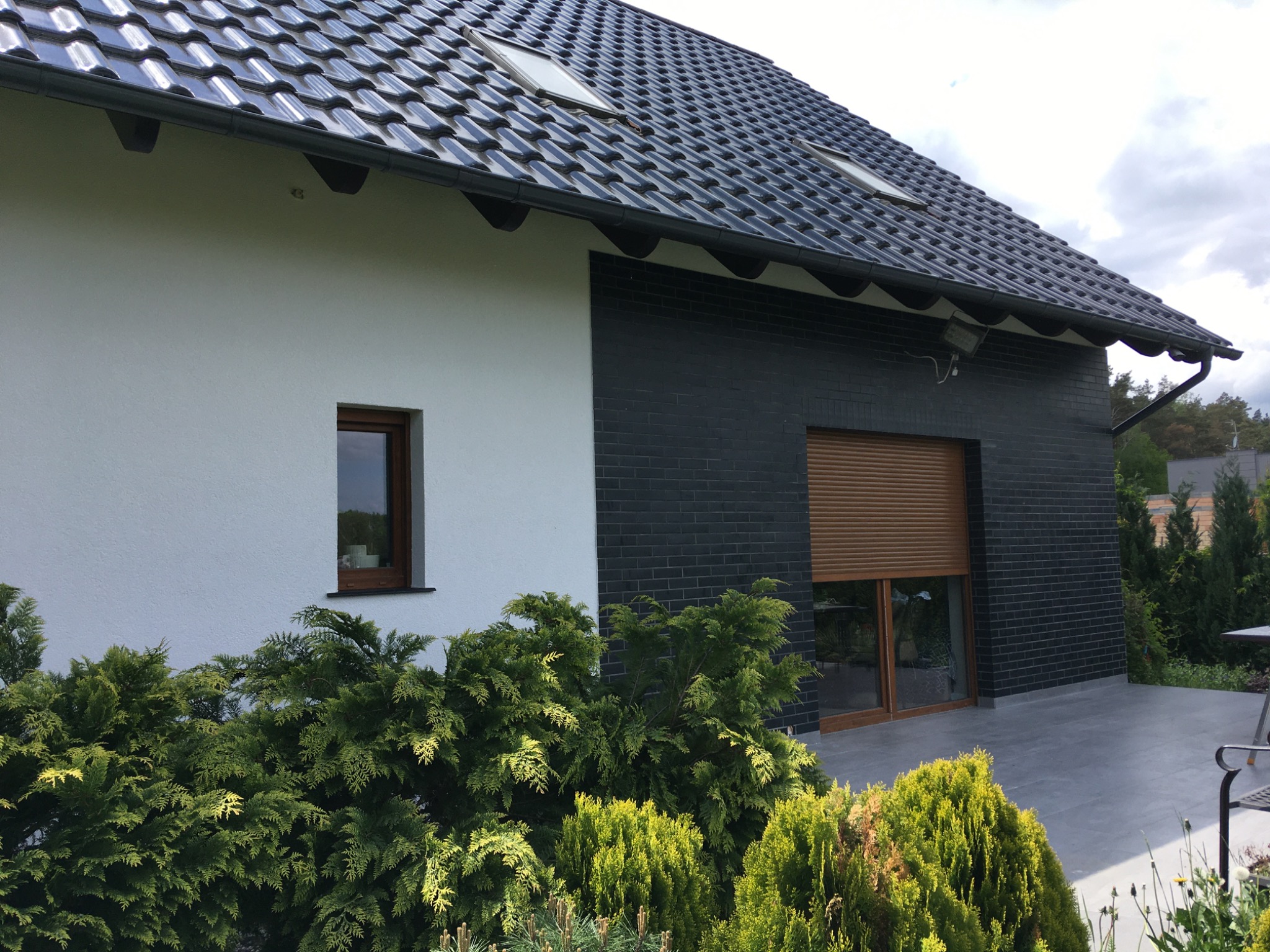 Nowoczesny dom z białą i grafitową elewacją, czarnym dachem z lukarnami i szarym tarasem, otoczony zielenią.