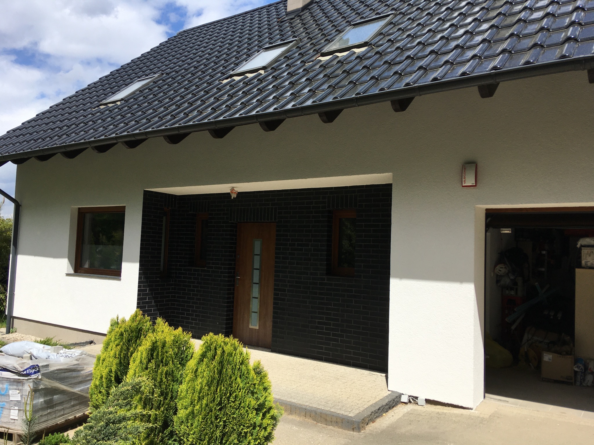 Wykończony dom jednorodzinny z czarną dachówką, białymi i czarnymi elewacjami, oraz widocznymi oknami dachowymi i zagospodarowanym terenem przed wejściem.