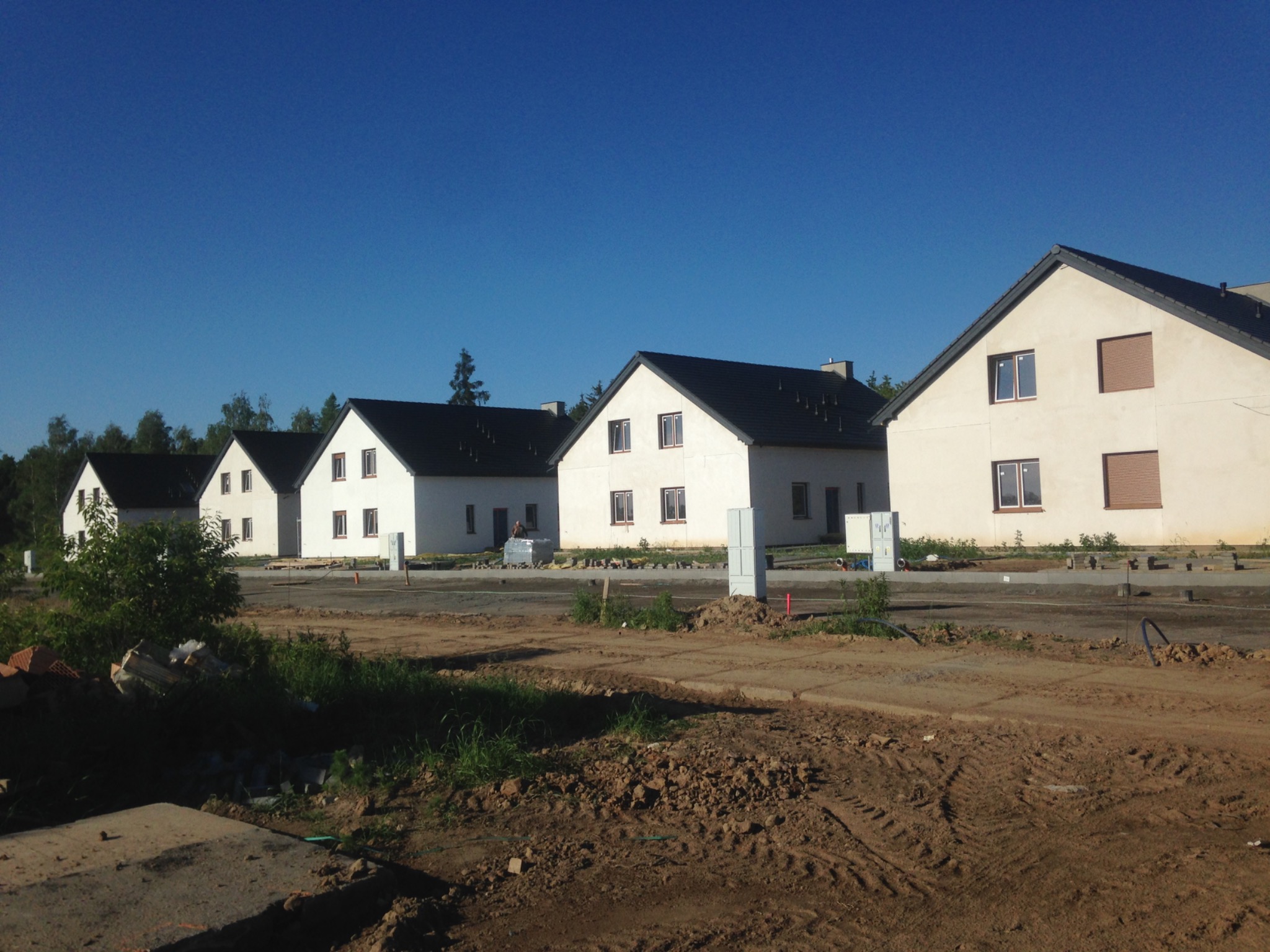 Rząd nowych, dwukondygnacyjnych domów z ciemnymi dachami i jasnymi elewacjami, w trakcie budowy, widoczny plac budowy i materiały budowlane.