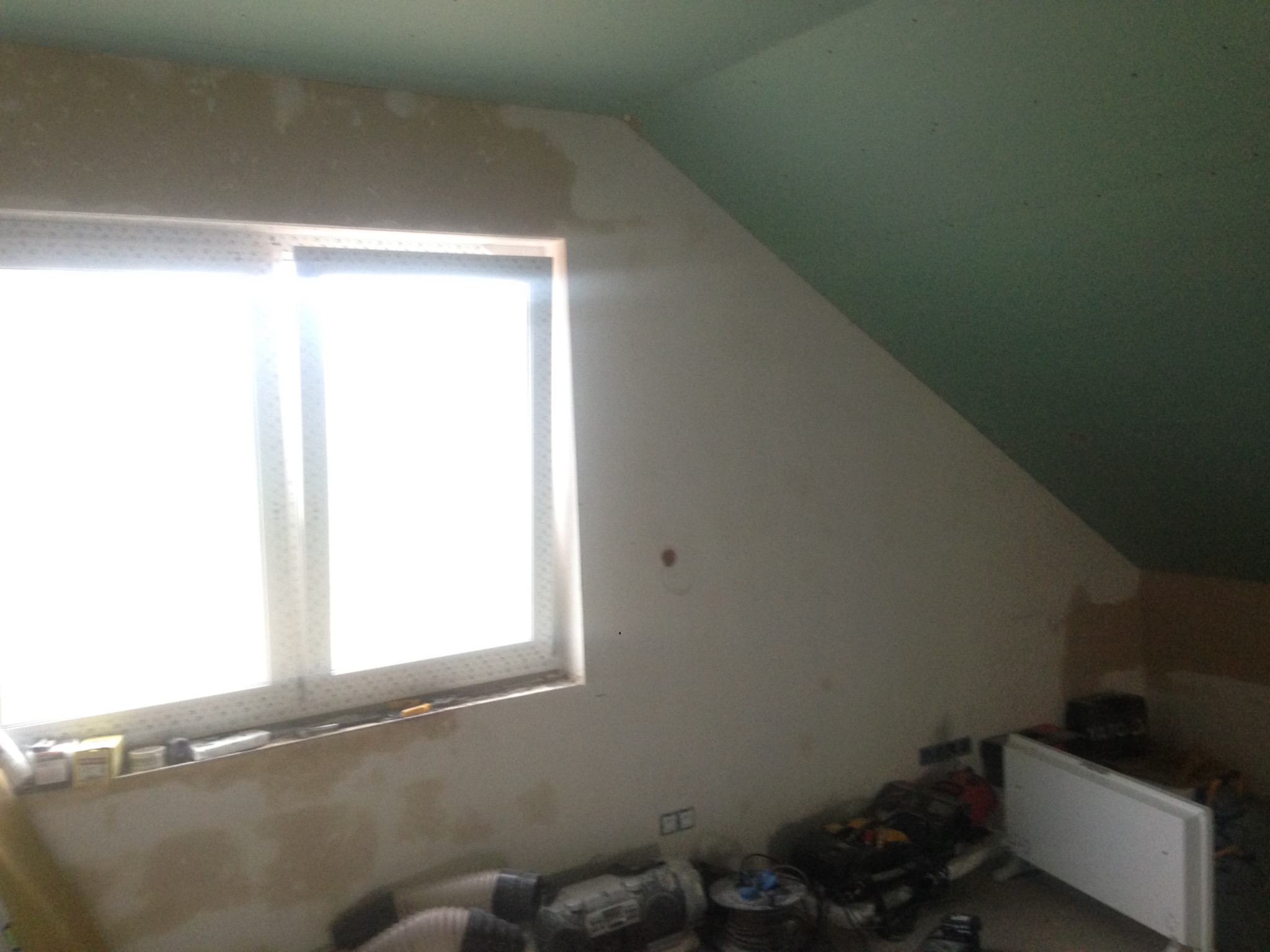 Poddasze w Rogoźnie w trakcie prac przygotowawczych, widoczne ślady po szpachlowaniu przy oknie, sufit częściowo pomalowany na zielono, na podłodze leżą narzędzia i materiały budowlane.