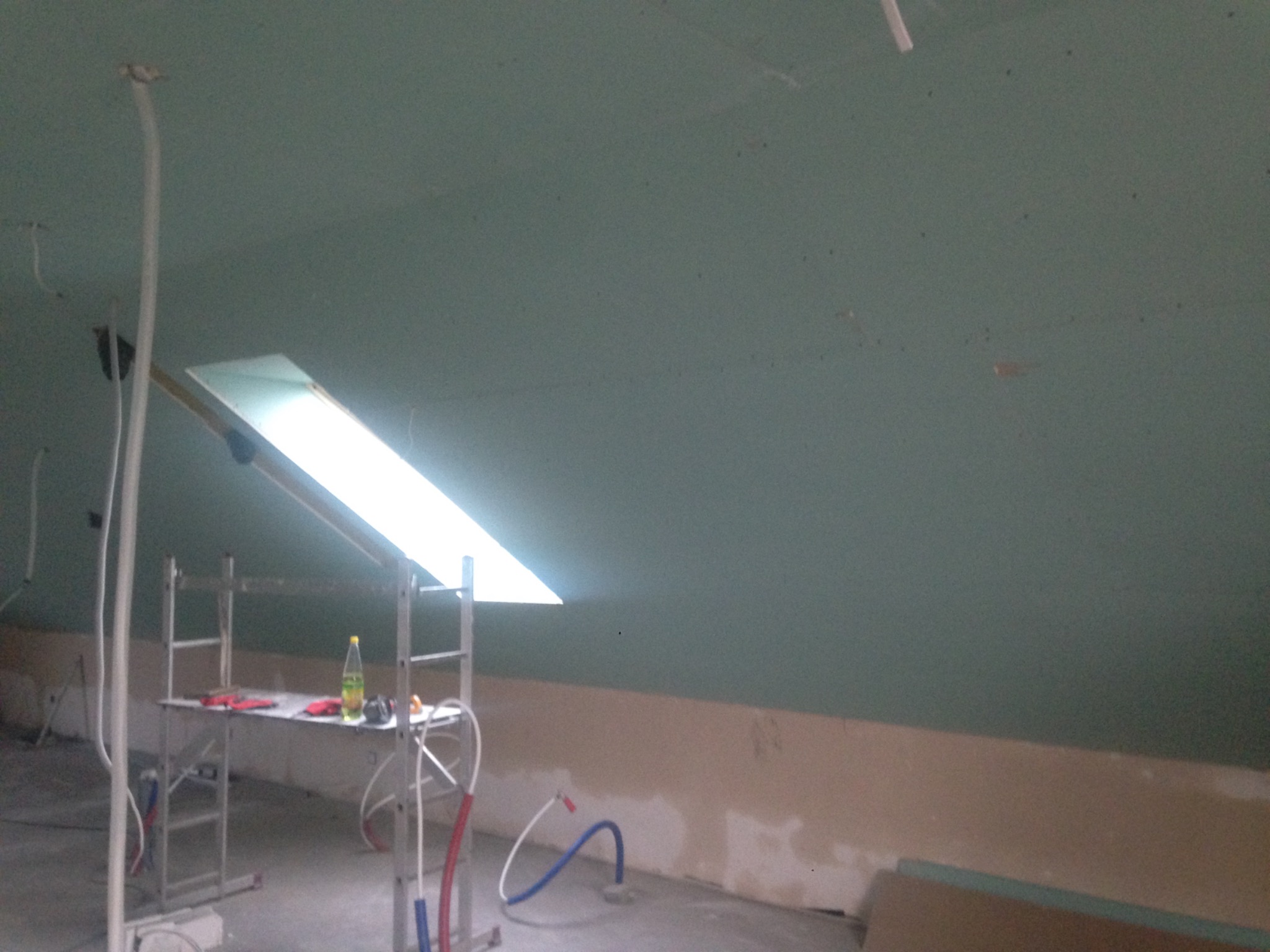 Poddasze w trakcie prac wykończeniowych: ściany pokryte płytami gipsowo-kartonowymi, widoczny świetlik dachowy, rusztowanie z narzędziami i butelką wody, wystające rury instalacyjne.