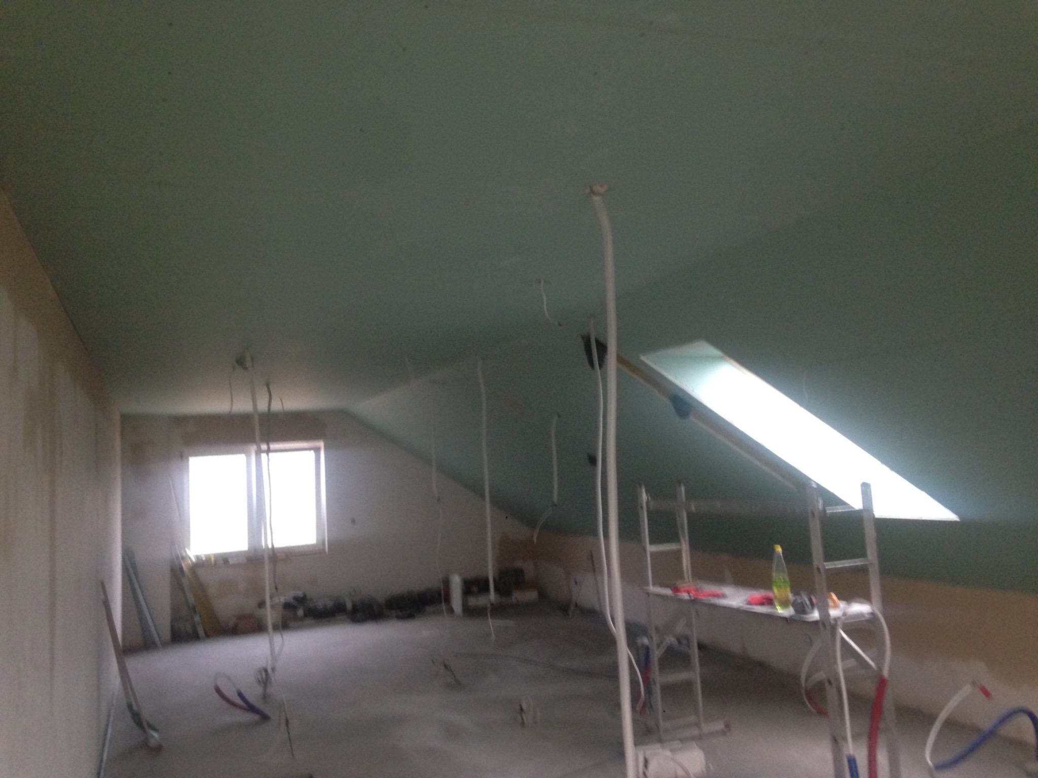 Poddasze w trakcie prac wykończeniowych: widoczne płyty kartonowo-gipsowe na skosach, okno dachowe, kable zwisające z sufitu i rusztowanie z narzędziami.
