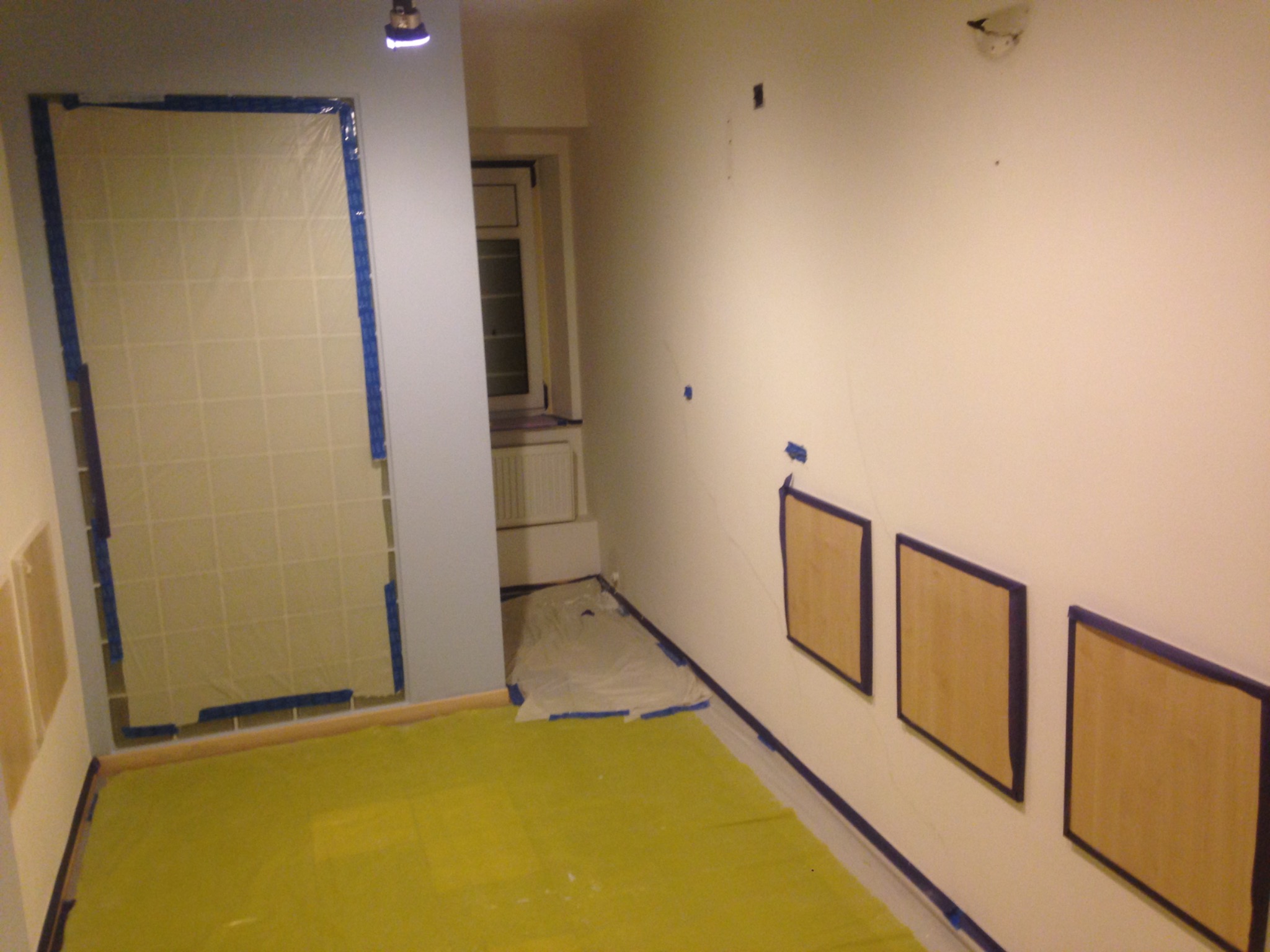 Pokój przygotowany do prac wykończeniowych: podłoga i drzwi zabezpieczone folią ochronną, ściany z zamaskowanymi ramami, widoczne ślady po poprzednich pracach.