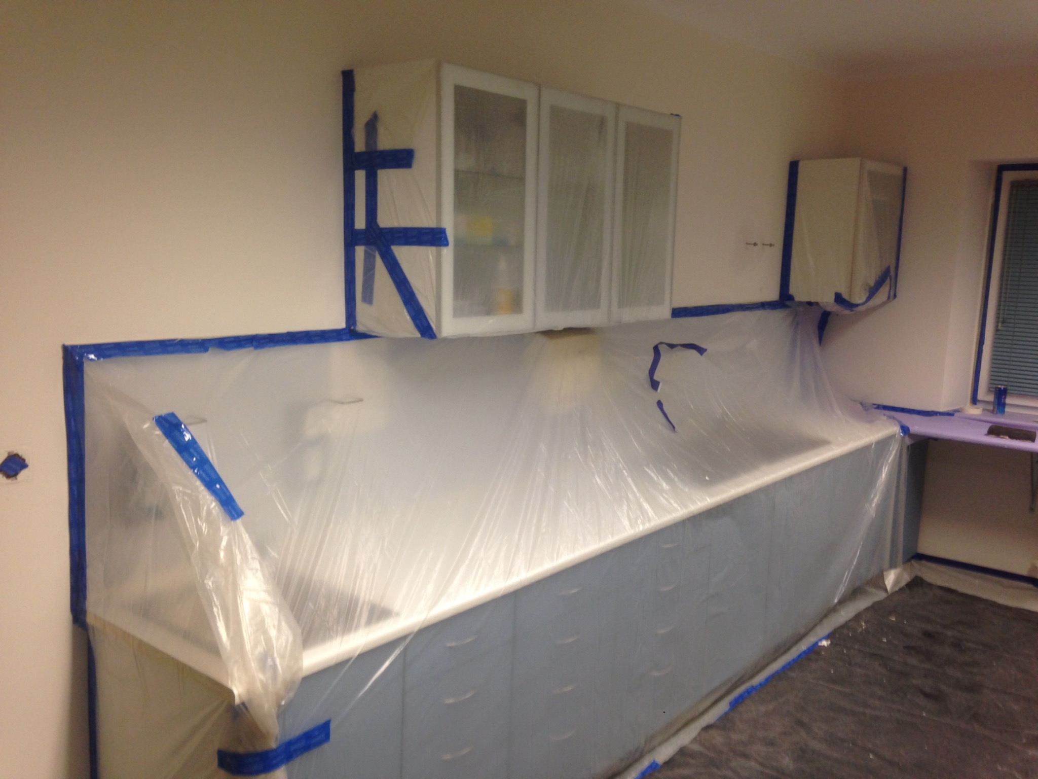 Przygotowanie łazienki do prac malarskich: meble i powierzchnie zabezpieczone folią i taśmą malarską, widoczne szafki wiszące, blat i fragment okna.