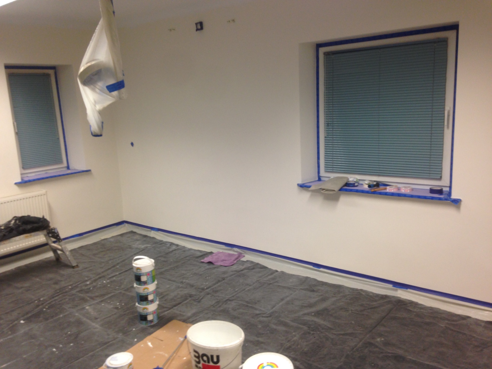 Przygotowane pomieszczenie do prac malarskich: zabezpieczone okna i listwy taśmą malarską, rozłożona folia ochronna na podłodze, widoczne wiadra z farbą i drabina.