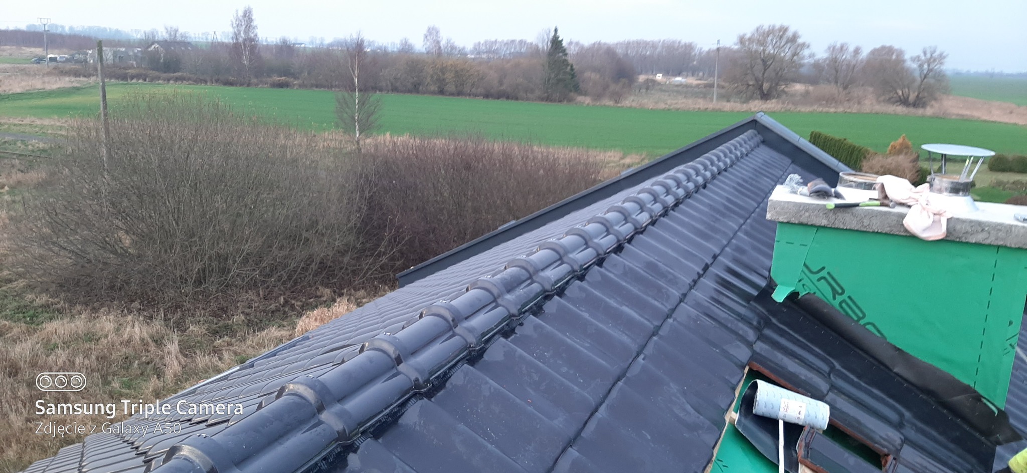 Czarny dach z dachówki ceramicznej widziany z góry, komin zabezpieczony zieloną folią, w tle pole i drzewa.