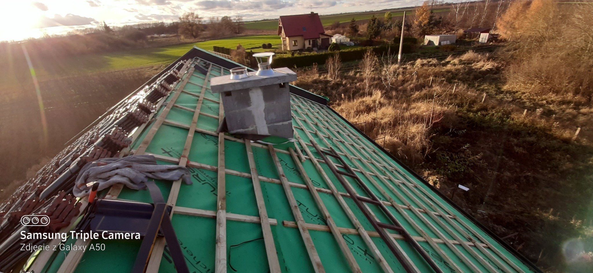 Montaż łat dachowych i membrany w Świerczu, widoczny komin i częściowo ułożona dachówka, w tle budynek mieszkalny.