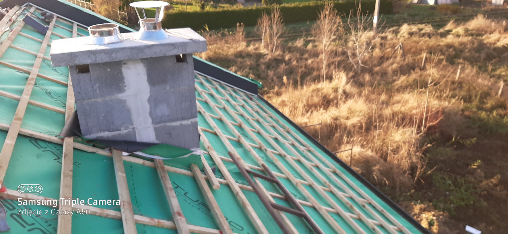 Dach w trakcie budowy z widocznym kominem z szarej cegły, pokryty zieloną membraną i drewnianą konstrukcją łat, w tle jesienny krajobraz.