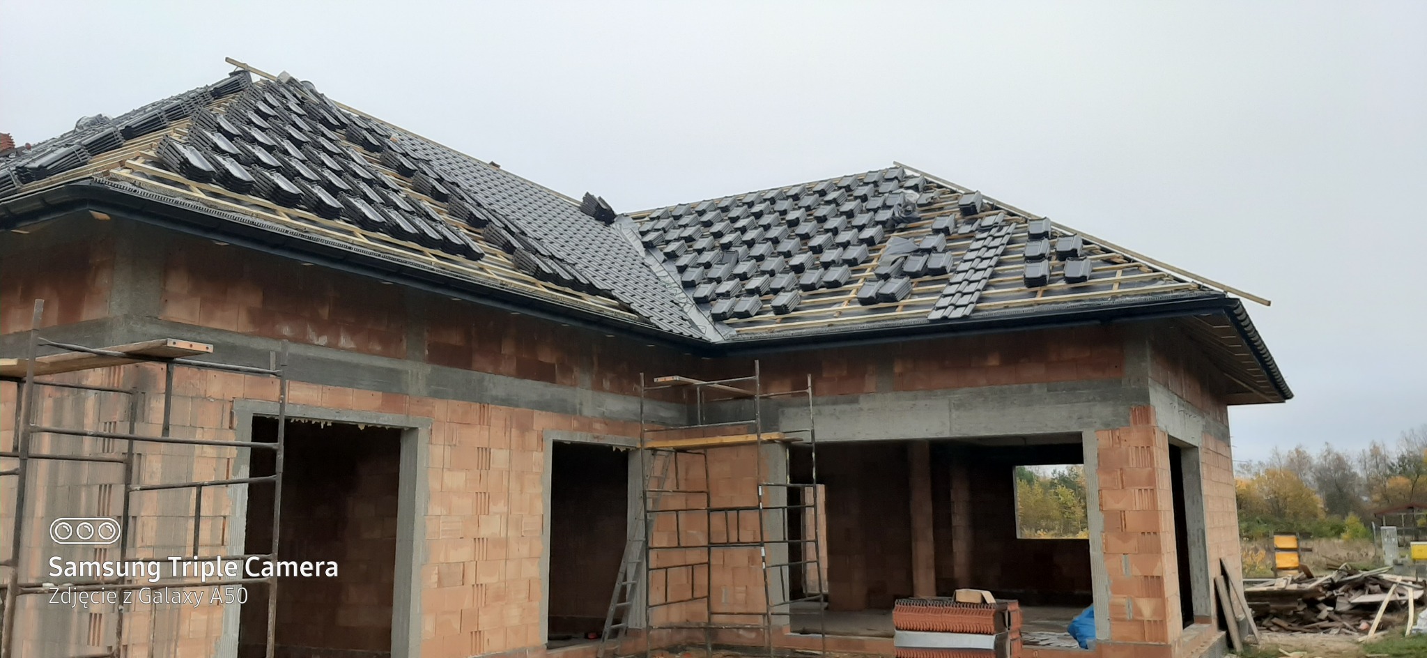 Dom w trakcie budowy z częściowo ułożonym, czarnym dachem ceramicznym. Widoczne drewniane łaty, rynna, oraz ściany z czerwonej cegły. Na pierwszym planie rusztowanie.