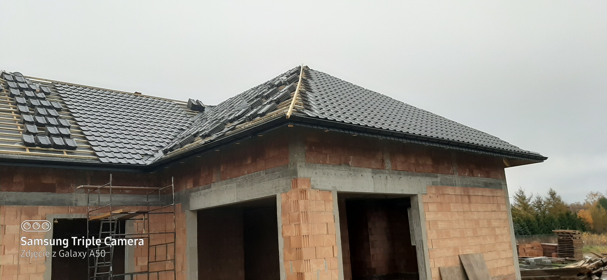 W trakcie układania czarnej dachówki ceramicznej na nowym dachu domu w stanie surowym otwartym, widoczne drewniane łaty i fragment zamontowanej rynny.