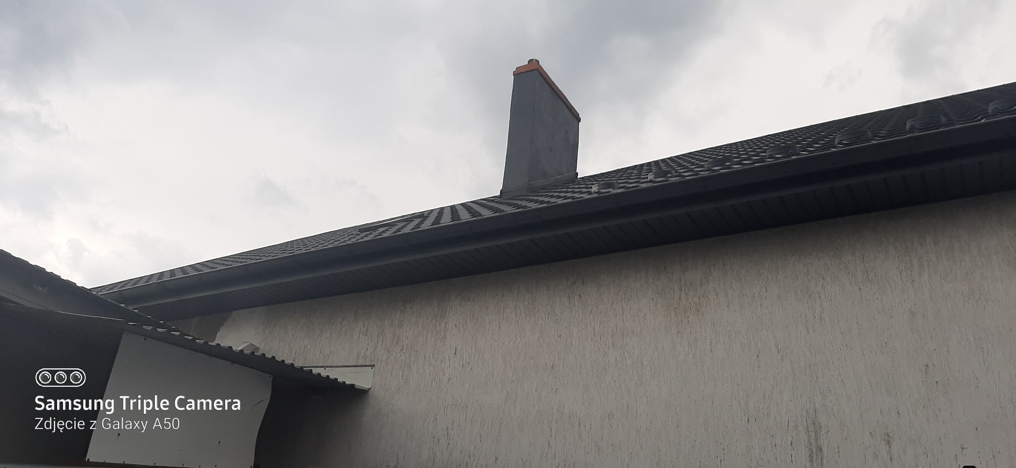 Ciemny dach z dachówki ceramicznej, szara rynna i komin z pomarańczowym daszkiem na tle pochmurnego nieba, widok z dołu.