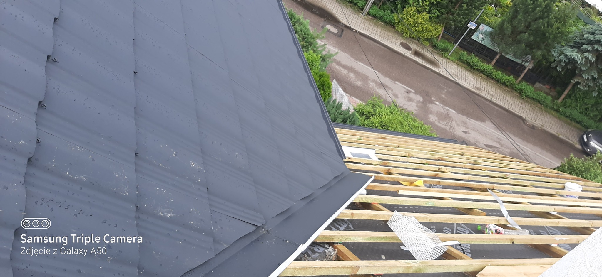Metalowy dach w trakcie montażu, widoczne krople deszczu na powierzchni, drewniana konstrukcja więźby dachowej, tło z zielenią i fragmentem ulicy.