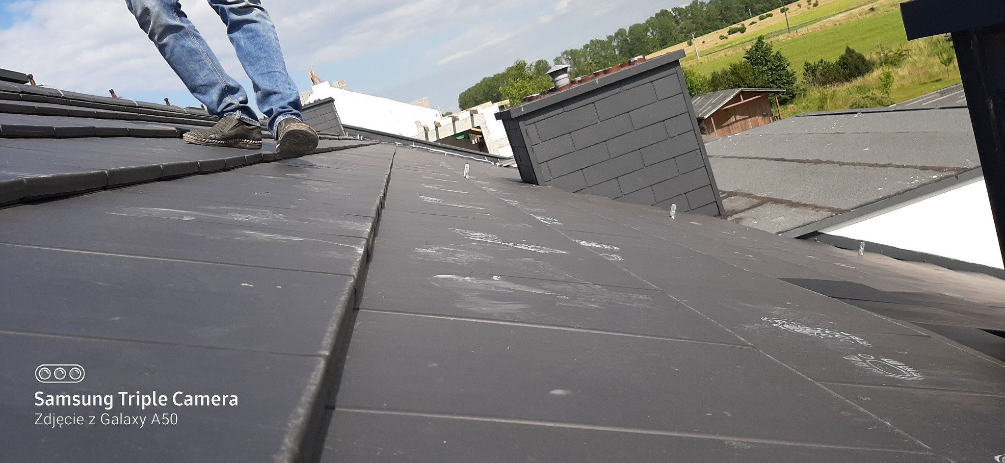 Pracownik w jeansach i ciemnych butach stoi na nowym, grafitowym dachu z dachówki ceramicznej w trakcie prac wykończeniowych, widoczny komin i fragmenty innych budynków w tle.