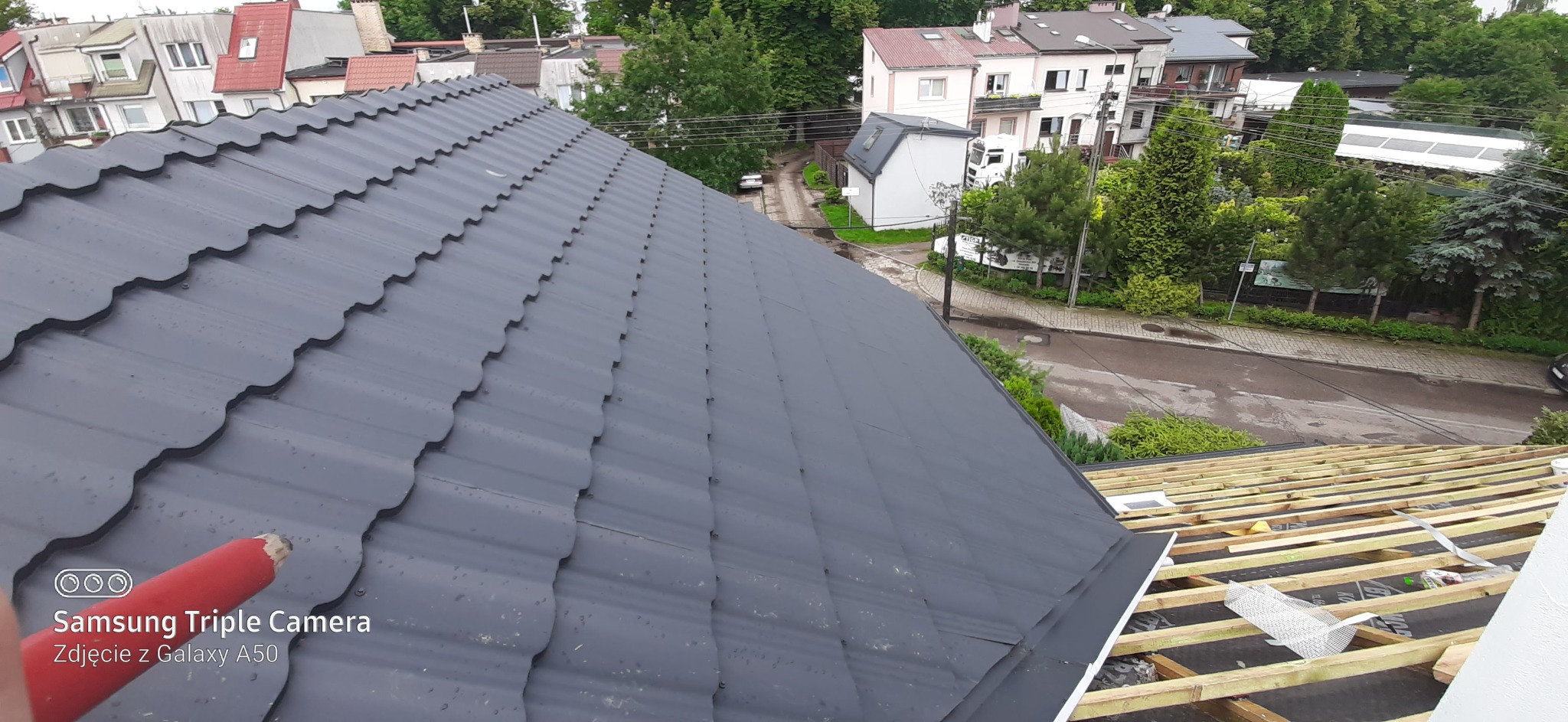 Ciemnoszara blachodachówka ułożona na dachu domu jednorodzinnego, widok z góry na fragment nowej konstrukcji więźby dachowej w tle zabudowa miejska i zieleń.