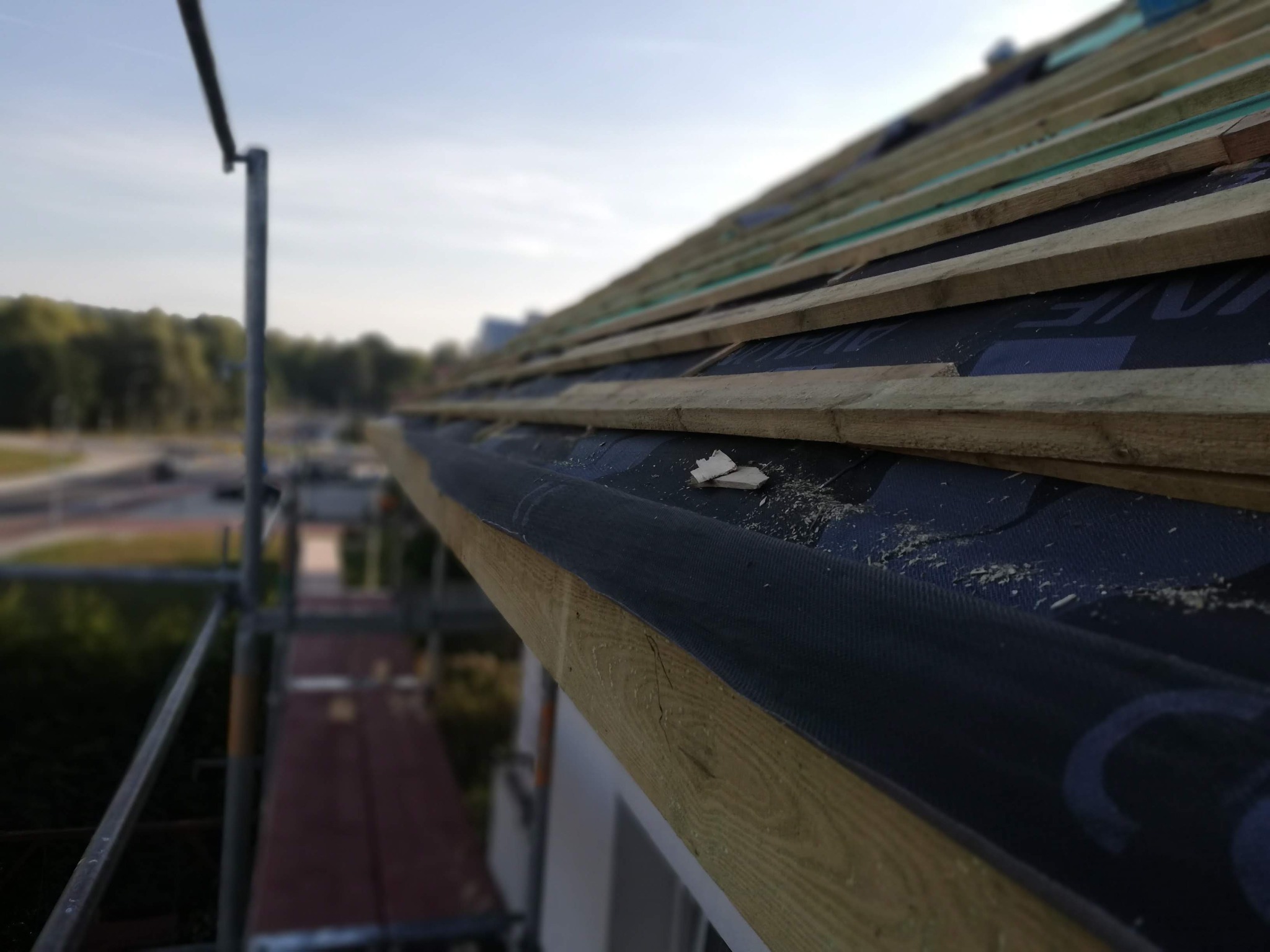 Konstrukcja dachu w trakcie budowy: widoczne drewniane krokwie, czarna membrana dachowa z delikatnym wzorem, fragment rusztowania w tle.