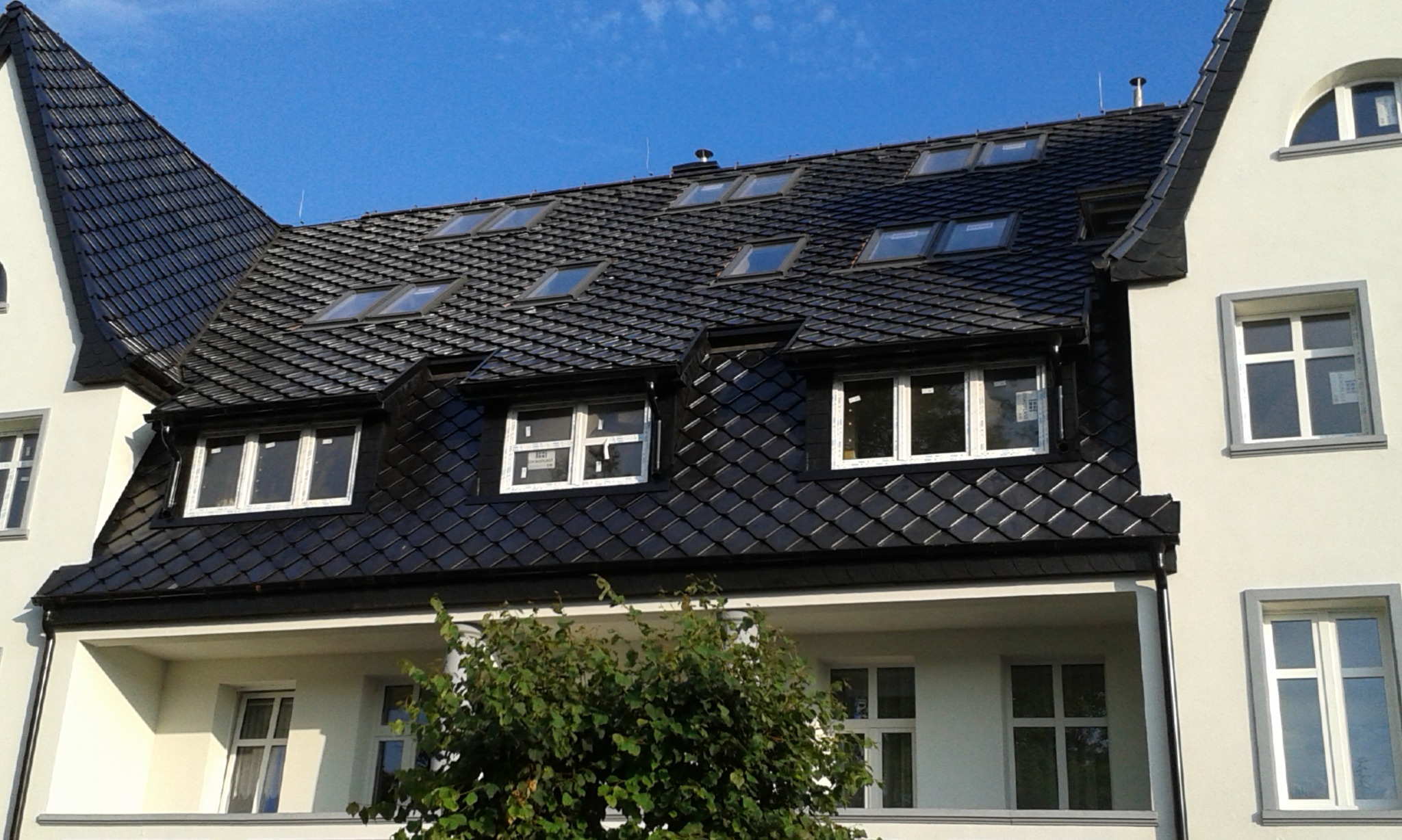 Nowoczesny budynek z ciemnym dachem pokrytym dachówką karpiówką i oknami dachowymi, widok z dołu na tle błękitnego nieba.