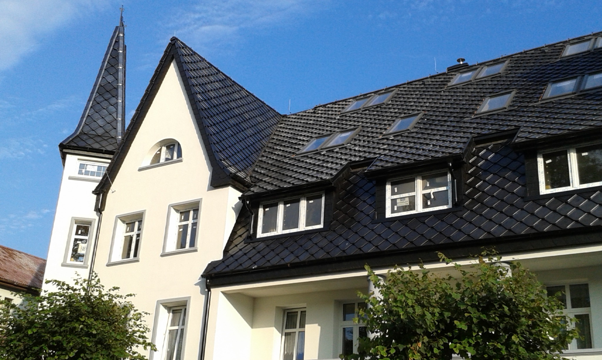 Elewacja budynku z czarnym dachem z dachówki łuskowej i wieżą pokrytą tym samym materiałem, widoczne okna dachowe i roślinność.