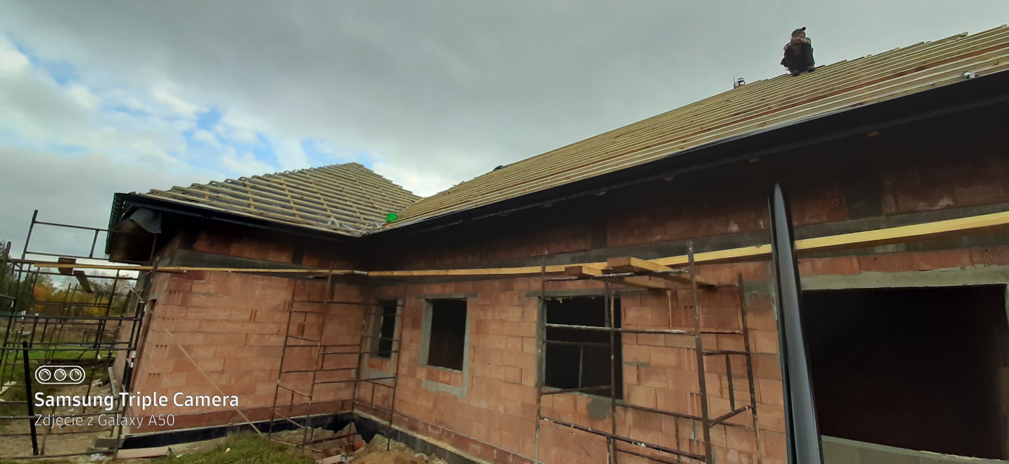 Budowa dachu jednopiętrowego domu z czerwonej cegły, widoczne drewniane łaty pod pokrycie dachowe, fragmenty rynien i pracownik na dachu, pochmurne niebo w tle.