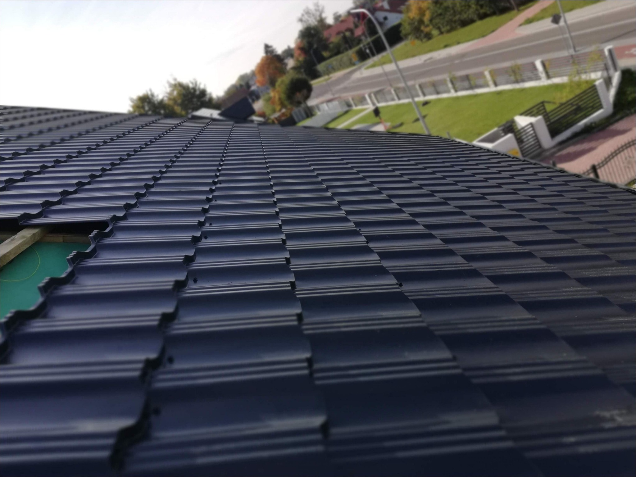 Nowy, ciemny dach z blachodachówki modułowej, widok z góry pod kątem, fragment deskowania widoczny w lewym górnym rogu, w tle zabudowa jednorodzinna.
