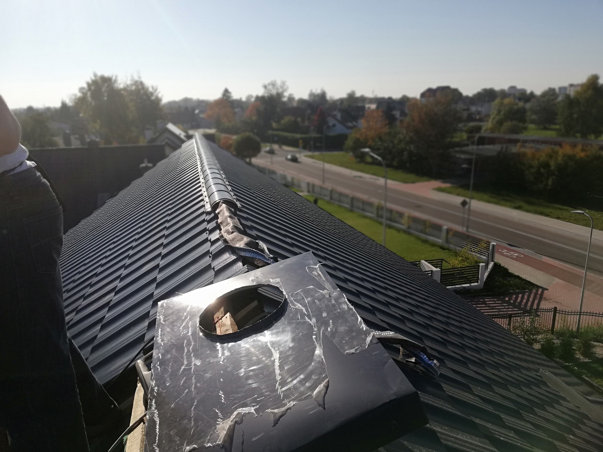 Czarny dach z blachy trapezowej w trakcie montażu z widocznym otworem na komin i częściowo usuniętą folią ochronną, w tle zabudowa miejska i droga.