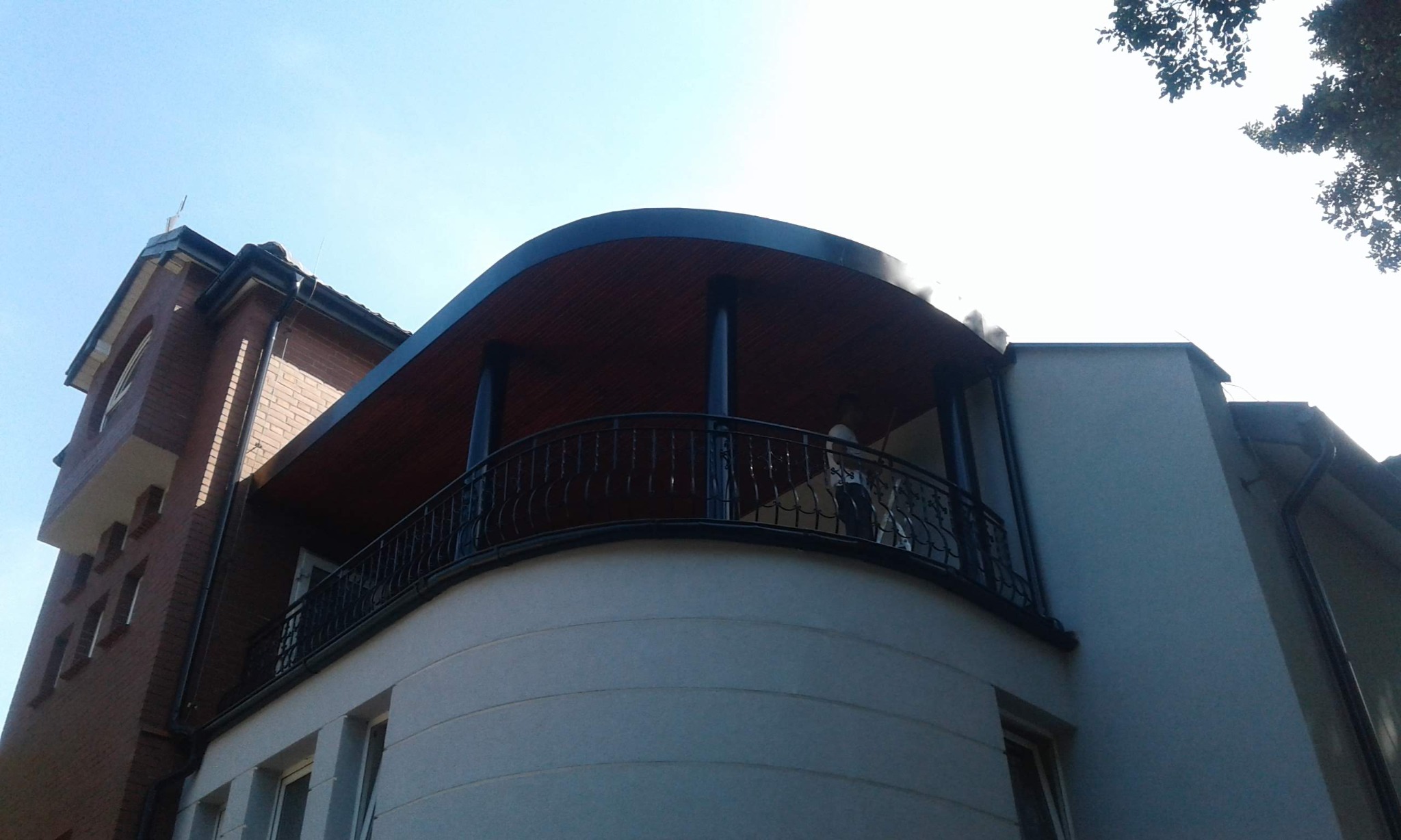 Widok z dołu na dwupiętrowy budynek z cegły i tynkowaną elewacją, z zaokrąglonym balkonem z czarną, kutą balustradą i drewnianym zadaszeniem w kolorze wiśniowym, z widocznymi rynnami.