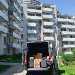 Otwarty tył szarego furgonetki z ładunkiem kartonów i walizek na tle nowoczesnego budynku mieszkalnego z balkonami i wieżowca Capital Towers w tle.