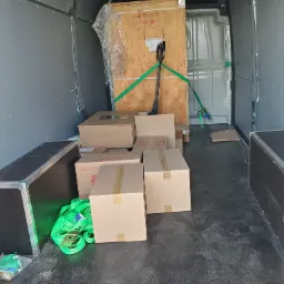 Wnętrze furgonetki wypełnionej kartonami i dużą drewnianą skrzynią zabezpieczoną zielonymi pasami transportowymi, widok z tyłu pojazdu.