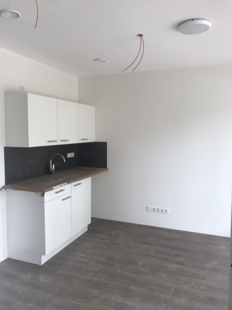 Wnętrze kuchni w stanie deweloperskim z widocznymi wystającymi przewodami elektrycznymi z sufitu, sugerującymi przyszłe oświetlenie, oraz zamontowanymi białymi szafkami kuchennymi z szarym blatem...