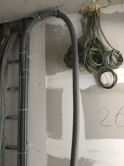 Szare kable elektryczne prowadzone w rurze ochronnej, mocowane do sufitu, z widoczną puszką przyłączeniową i zwisającymi przewodami na tle surowej ściany w trakcie prac wykończeniowych.