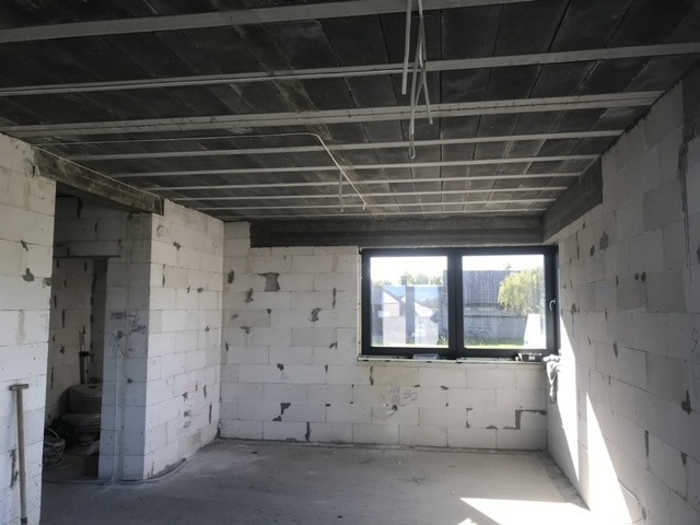 Surowe wnętrze domu w trakcie budowy z widocznymi bloczkami z betonu komórkowego, żelbetowym stropem, oknem z czarną ramą i przewodami elektrycznymi zwisającymi z sufitu.