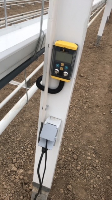 Żółto-czarny miernik przymocowany do białej, metalowej konstrukcji, z widoczną puszką przyłączeniową i przewodami na tle ziemi.