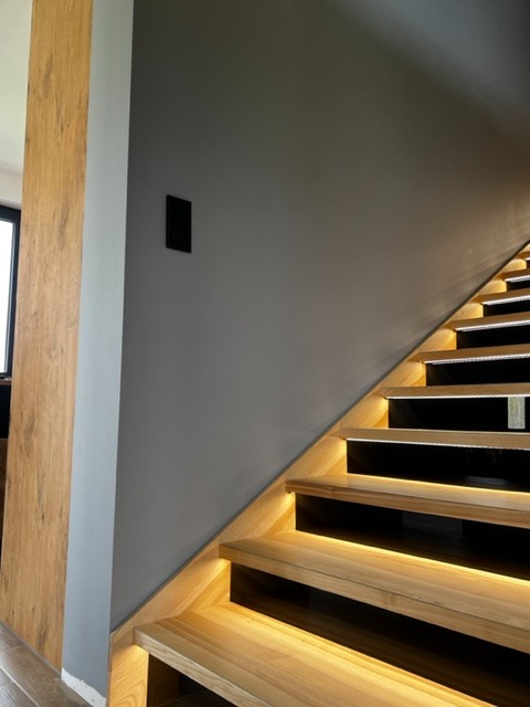 Oświetlenie LED schodowe z drewnianymi stopniami i czarnymi podstopnicami, widoczny włącznik na szarej ścianie.