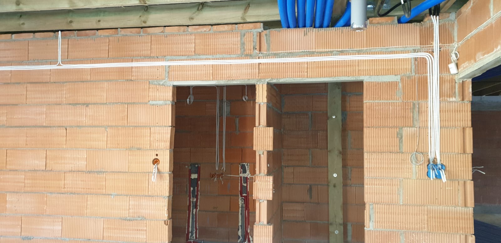 Surowy stan budynku w trakcie prac: widoczne przewody elektryczne prowadzone po ścianach z cegły, instalacja podtynkowa w otworach drzwiowych, niebieskie rury instalacyjne pod stropem.
