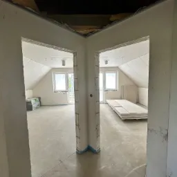 Dwa pokoje na poddaszu w trakcie prac wykończeniowych, widoczne surowe ściany, okna dachowe, płyty gipsowe i materiały budowlane, zarysowane ściany działowe z widoczną konstrukcją.