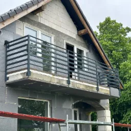 Dom w trakcie ocieplania styropianem grafitowym, z zamontowaną czarną, metalową balustradą balkonową i widocznym fragmentem rusztowania.