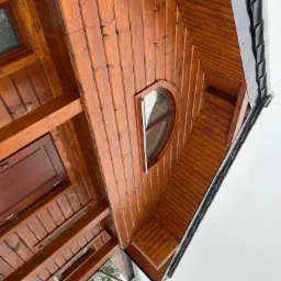 Drewniana elewacja domu z oknem w kształcie elipsy, widok od dołu, fragment dachu pokryty czarną blachodachówką.