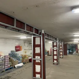 Stelaż z profili stalowych i płyt w kolorze bordo, prawdopodobnie przygotowany pod montaż płyt kartonowo-gipsowych, w surowym wnętrzu magazynu z zapasami napojów.