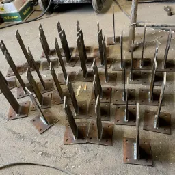 Przygotowane do montażu stalowe słupki z przyspawanymi podstawami, ułożone rzędami na betonowej posadzce warsztatu, widoczne ślady spawania i narzędzia w tle.