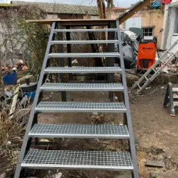 Stalowe schody z kratowymi stopniami w surowym otoczeniu budowy, widoczny spawarka Kemppi i drabina aluminiowa