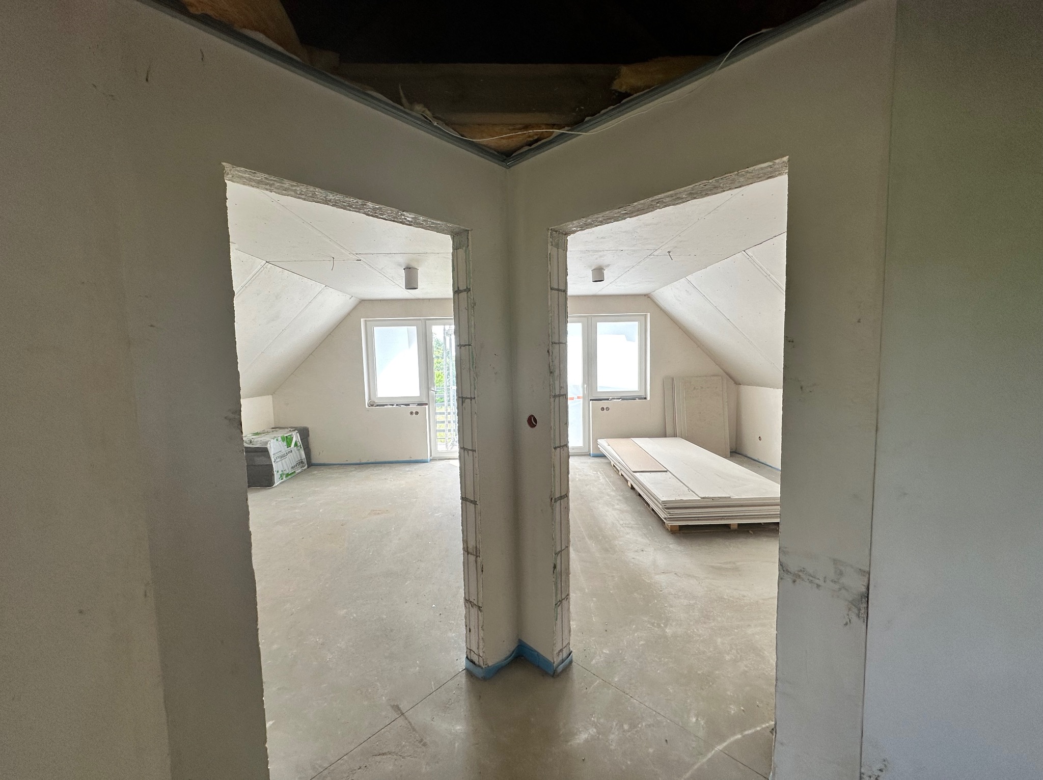 Dwa pokoje na poddaszu w trakcie prac wykończeniowych, widoczne surowe ściany, okna dachowe, płyty gipsowe i materiały budowlane, zarysowane ściany działowe z widoczną konstrukcją.