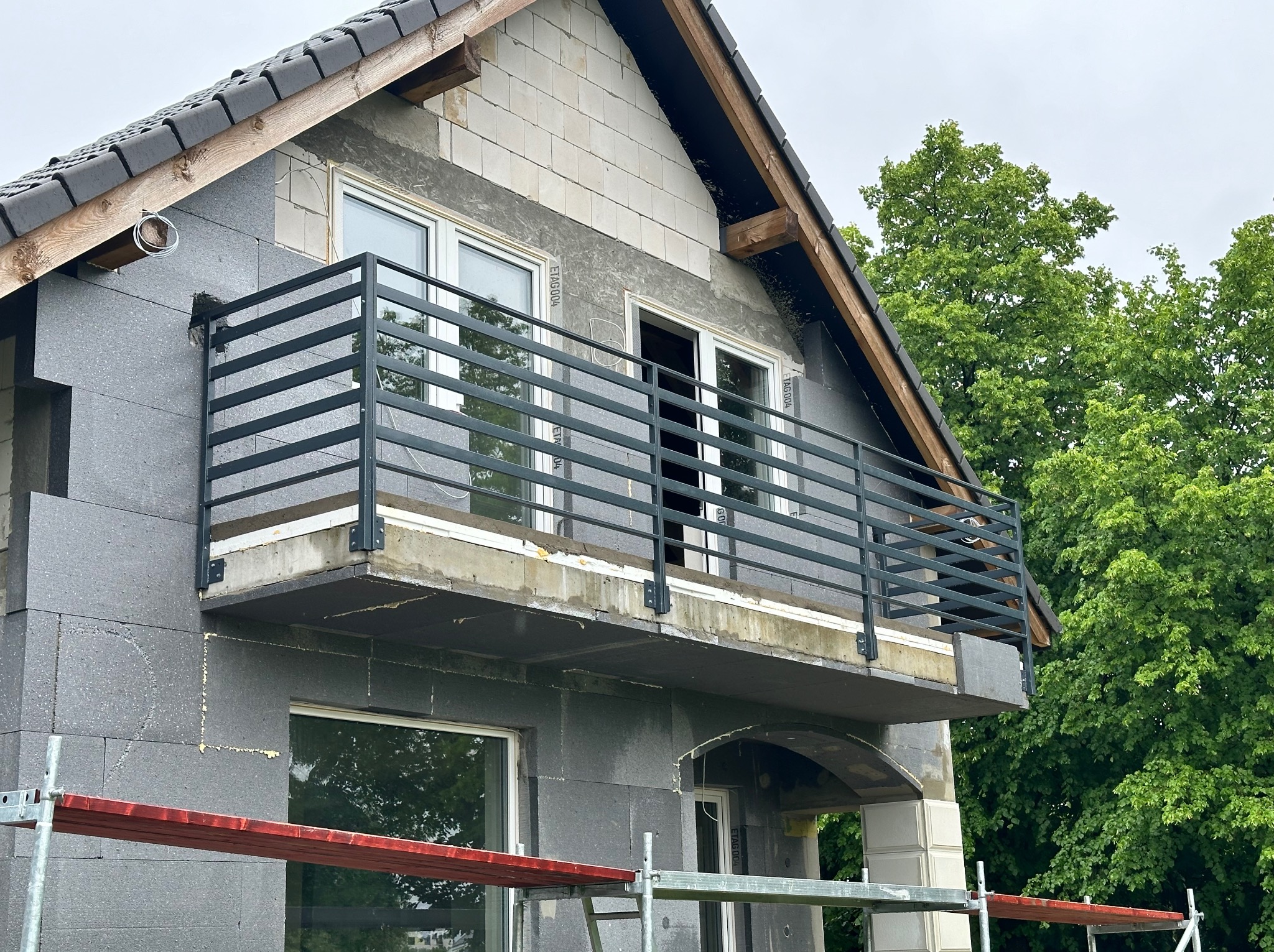 Dom w trakcie ocieplania styropianem grafitowym, z zamontowaną czarną, metalową balustradą balkonową i widocznym fragmentem rusztowania.